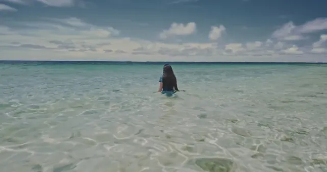 Image d'un film où l'on voit la poétesse Kathy Jetñil-Kijiner. Elle est de dos, les cheveux long relâchés, habillée, dans l'eau transparente de la mer qui lui arrive à la taille.