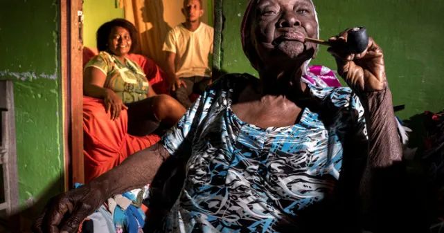 Une vieille femme brésilienne fume la pipe. On voit sa famille en arrière-plan.