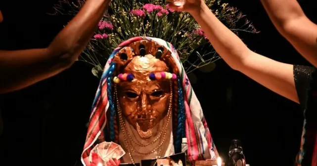 Trois bras devant un masque doré représentant un visage décoré de tissus, fleures et autres offrandes.